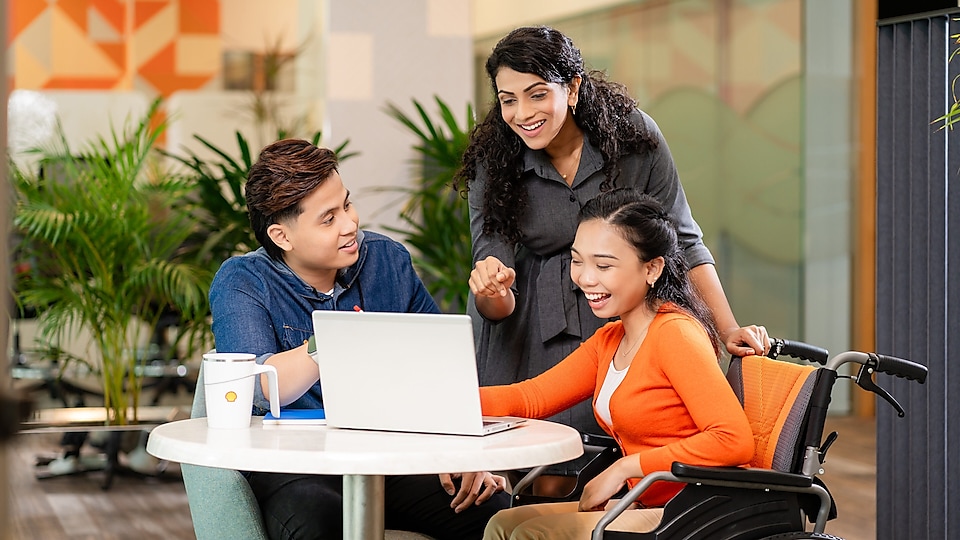 Three diverse people discussing around a laptop in a modern office. A woman in a wheelchair smiles while using the laptop, a man leans forward, and another woman stands behind, gesturing towards the screen. The background features plants and contemporary decor.