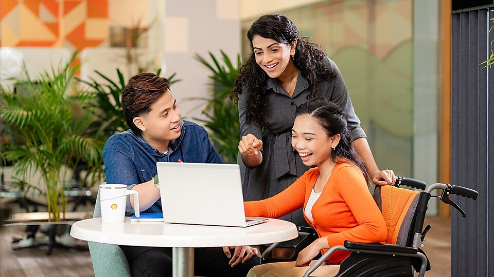 Three colleagues are gathered around a laptop, sharing a candid moment and smiling. The employee in the middle is standing, while the two others are seated, with one on a wheeled chair.